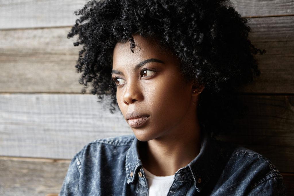 Black woman wearing denim shirt, in front of wood clad wall