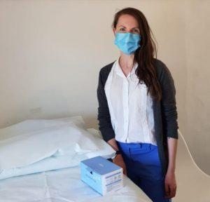 Helen standing in clinic wearing mask, grey cardigan, white shirt, blue trousers