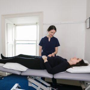 patient on treatment bed with acupuncturist standing at side. White wall and window background